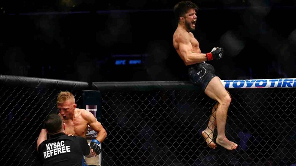 Henry Cejudo (atop cage) defeats TJ Dillashaw to remain flyweight champion, in 2019